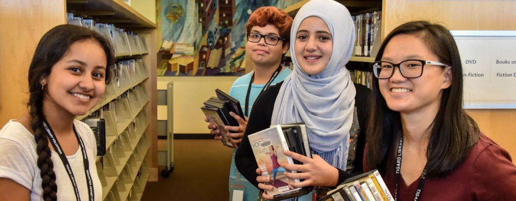 Four diverse teens holding DVDs and books in a library aisle filled with shelves and labeled sections.