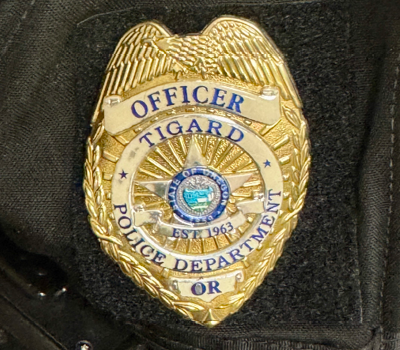 A close-up image of a gold Tigard Police Department badge with blue lettering, an eagle at the top, and a circular center emblem. The badge includes the words 'Officer,' 'Tigard Police Department,' and 'OR,' along with an established date.