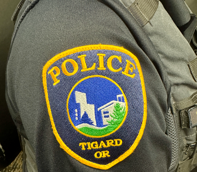 A close-up of a police uniform shoulder patch. The patch is blue with yellow trim and lettering, reading 'POLICE TIGARD OR,' and features a graphic of buildings and greenery.